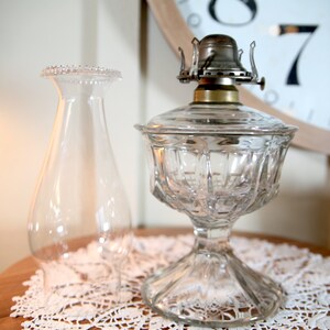 May include: A clear glass oil lamp with a decorative, faceted base and a clear glass chimney. The lamp has a silver-toned metal burner and a brass collar.