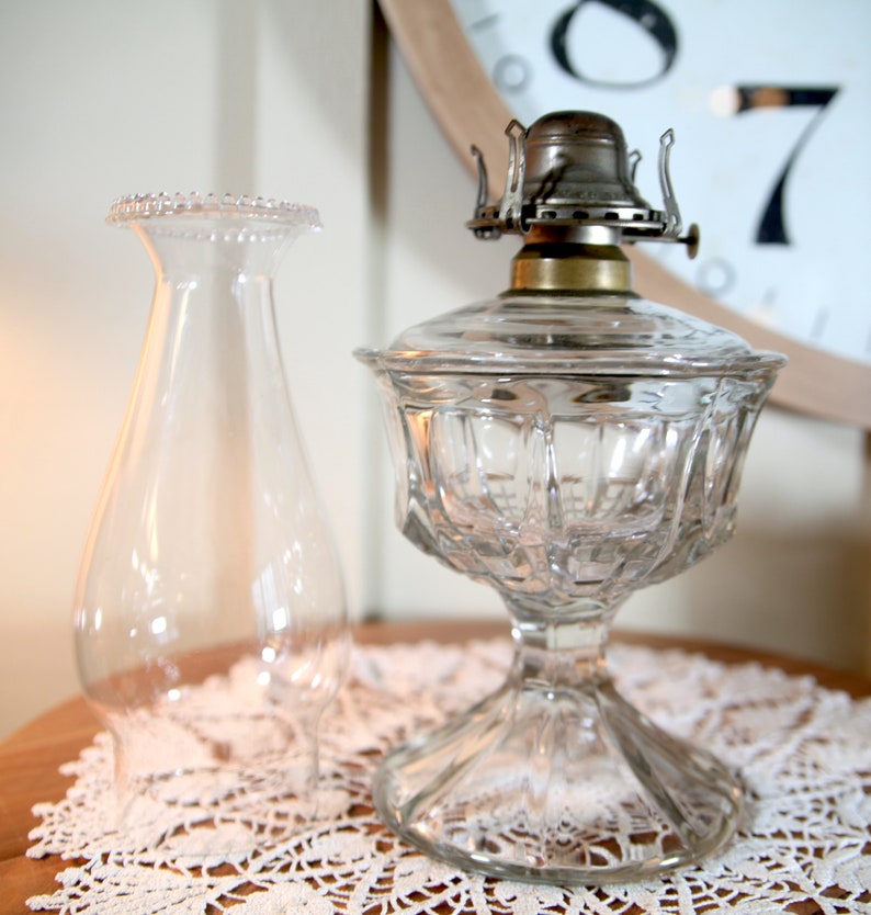 May include: A clear glass oil lamp with a decorative, faceted base and a clear glass chimney. The lamp has a silver-toned metal burner and a brass collar.