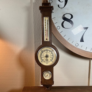 Puede incluir: Una estación meteorológica de madera con un barómetro, un termómetro y un higrómetro. El barómetro tiene una esfera blanca con marcas negras y el texto "RAIN - FAIR". El termómetro y el higrómetro tienen esferas blancas con marcas negras.