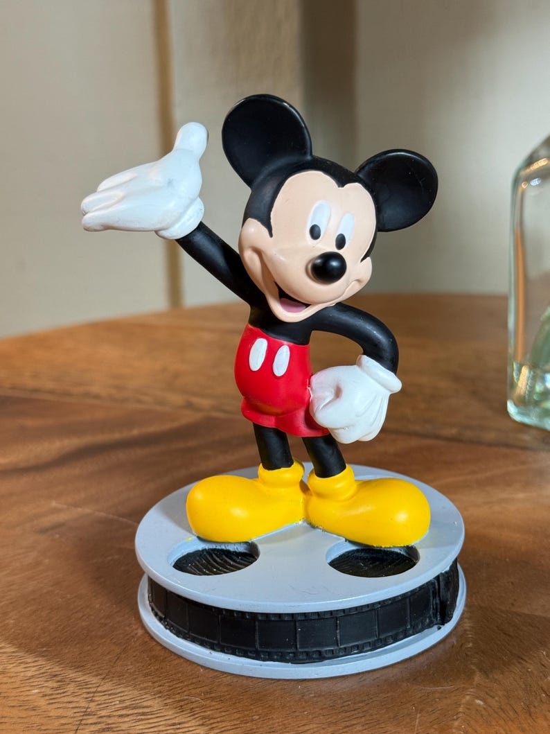 May include: A Mickey Mouse figurine, featuring black ears, a black body, red shorts, and yellow shoes. The figure is mounted on a grey film reel base. Mickey's white gloved hand is raised. A classic Disney collectible.