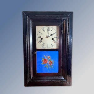 Puede incluir: Un reloj de pared rectangular con un marco de madera de color marrón oscuro. La sección superior presenta una esfera blanca con números romanos y manecillas negras. La sección inferior contiene un panel azul brillante con un motivo floral de rosas.