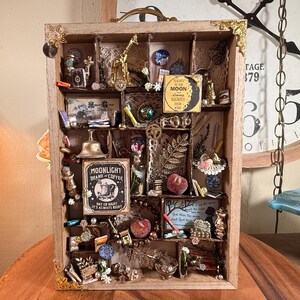 May include: A wooden shadow box with multiple compartments filled with miniature objects. The box contains a gold giraffe, a bell, a sign that says "MOONLIGHT BRAND COFFEE", and other small decorative items. The box has gold corner accents.
