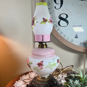 May include: A white and pink glass lamp with floral designs. The lamp has a white glass shade with pink and green floral accents. The base is a pink glass globe with a white floral design. The lamp has a brass base with a decorative design.