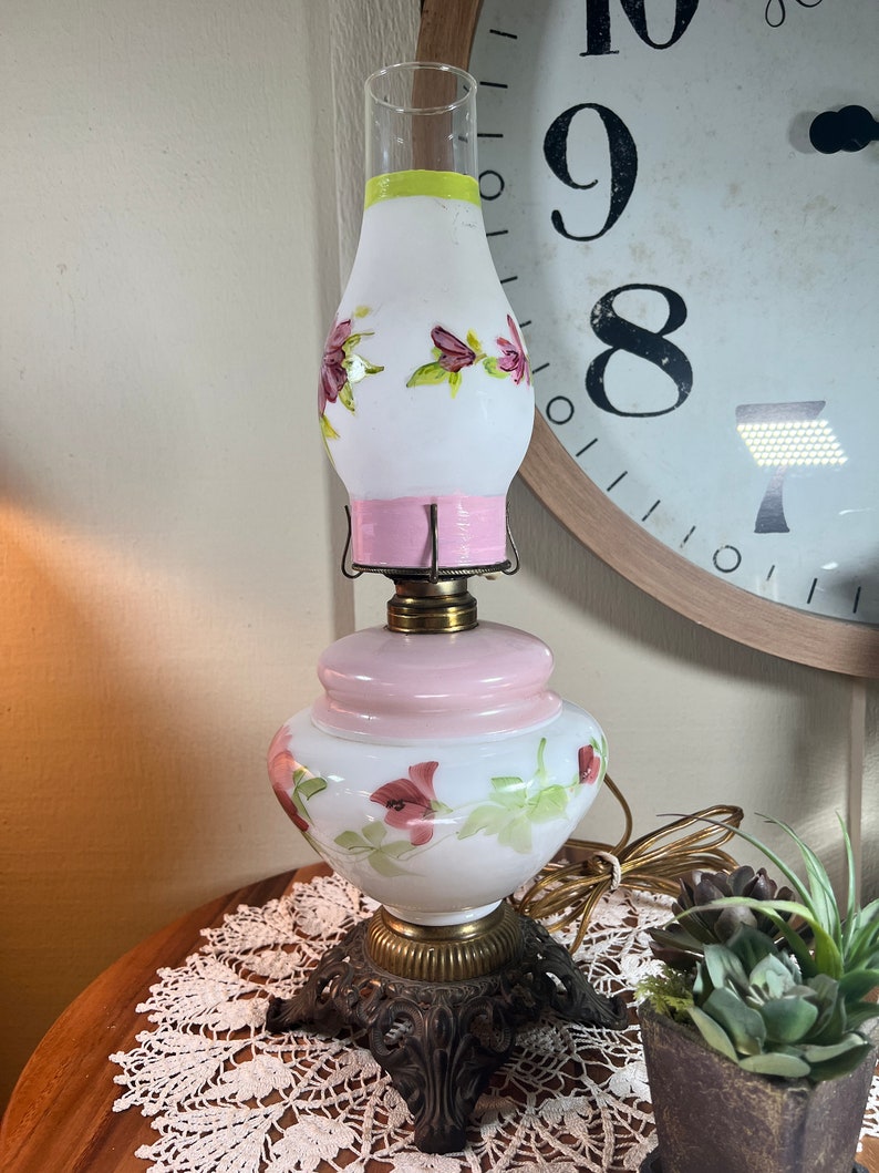 May include: A white and pink glass lamp with floral designs. The lamp has a white glass shade with pink and green floral accents. The base is a pink glass globe with a white floral design. The lamp has a brass base with a decorative design.
