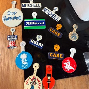 May include: A collection of vintage pin-back buttons and name tags with various designs and text. The buttons include phrases like "Stop Complaining" and "Freedom is Not Free." The name tags display names such as "Mitchell" and "Adlai."