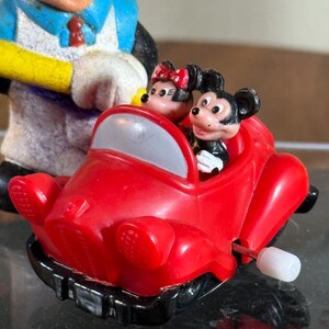 May include: A vintage Mickey Mouse and Minnie Mouse toy car. The red car has a white wind-up key and a gray windshield. Mickey Mouse is driving, and Minnie Mouse is in the passenger seat. A Mickey Mouse figurine is in the background.