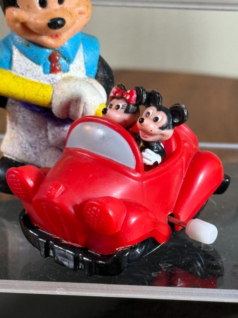 May include: A vintage Mickey Mouse and Minnie Mouse toy car. The red car has a white wind-up key and a gray windshield. Mickey Mouse is driving, and Minnie Mouse is in the passenger seat. A Mickey Mouse figurine is in the background.