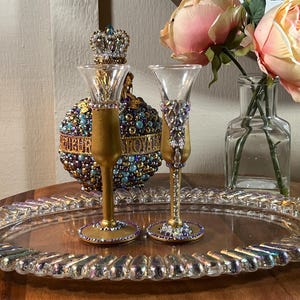 K&ouml;nnte beinhalten: Zwei goldbemalte Glasschnapsgl&auml;ser mit Juwelenbesatz auf einem durchsichtigen Tablett. Die Gl&auml;ser sind mit lila, blauen und goldenen Perlen verziert.