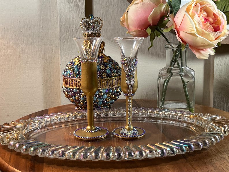 K&ouml;nnte beinhalten: Zwei goldbemalte Glasschnapsgl&auml;ser mit Juwelenbesatz auf einem durchsichtigen Tablett. Die Gl&auml;ser sind mit lila, blauen und goldenen Perlen verziert.