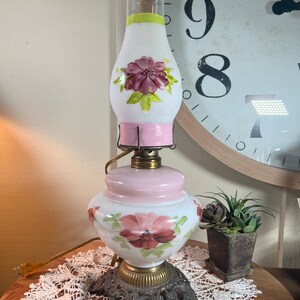 May include: A vintage white and pink glass lamp with floral designs. The lamp has a metal base with a decorative scroll design. The lamp is lit and the light shines through the glass.