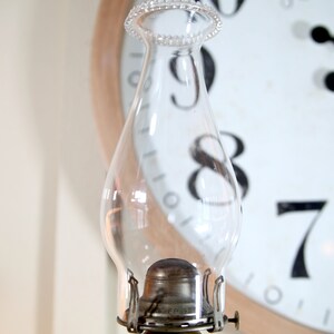May include: A clear glass oil lamp chimney with a metal base and a decorative beaded rim. The chimney is sitting in front of a clock face with Roman numerals.