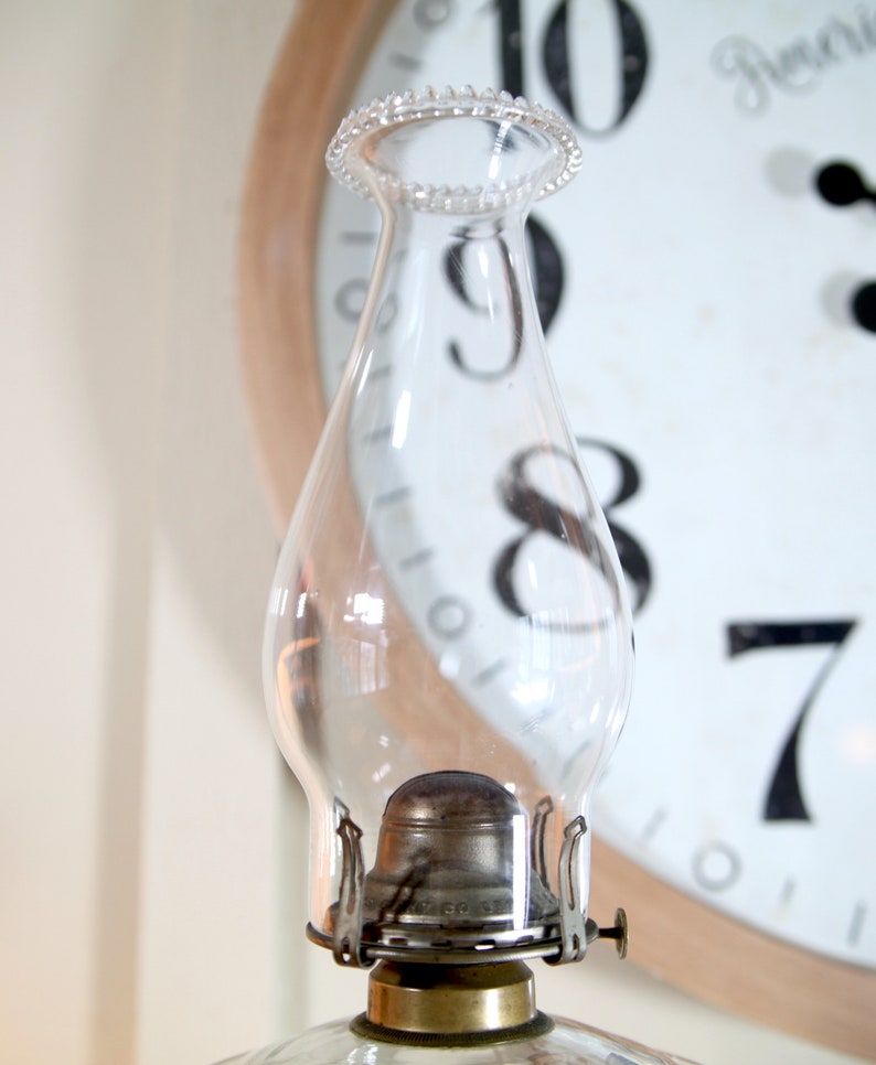 May include: A clear glass oil lamp chimney with a metal base and a decorative beaded rim. The chimney is sitting in front of a clock face with Roman numerals.