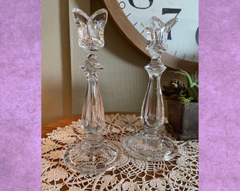 May include: Two clear glass candle holders with a tulip-shaped top and a faceted design. The candle holders are sitting on a white lace doily.