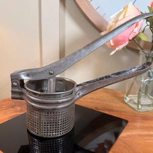 May include: An antique, metal potato ricer with a perforated bowl and long handles. The ricer is silver-toned and shows signs of wear, suggesting its age. It sits on a black surface, with a wooden table and a vase of flowers in the background.
