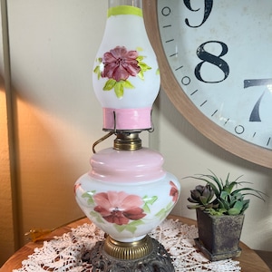 May include: A white glass lamp with a pink base and floral designs. The lamp has a white and green glass shade with a pink band around the top. The lamp is sitting on a white lace doily on a wooden table.
