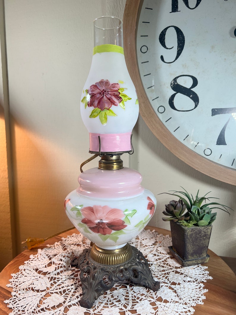 May include: A white glass lamp with a pink base and floral designs. The lamp has a white and green glass shade with a pink band around the top. The lamp is sitting on a white lace doily on a wooden table.
