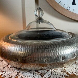May include: A silver hammered metal serving dish with a glass lid and a clear glass knob. The dish has a textured surface and a round shape.