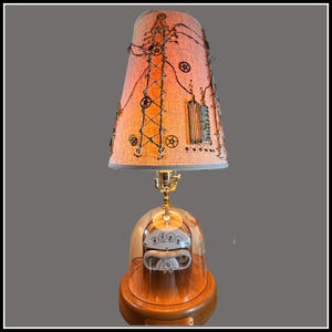 May include: A unique steampunk-style table lamp. The lamp features a peach-colored lampshade adorned with gears, wires, and a stylized power tower design. The base is a wooden dome with a glass cover, housing a vintage-looking gauge.