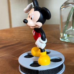 May include: A Mickey Mouse figurine in a classic pose, wearing red shorts, yellow shoes, white gloves, and a white hat. The figurine is standing on a grey film reel base. The figure is black, red, yellow, white, and tan.