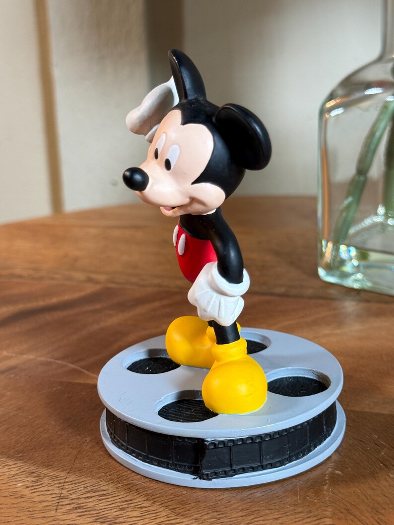 May include: A Mickey Mouse figurine in a classic pose, wearing red shorts, yellow shoes, white gloves, and a white hat. The figurine is standing on a grey film reel base. The figure is black, red, yellow, white, and tan.