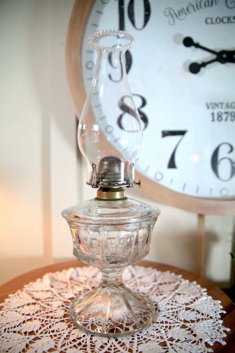 May include: A clear glass oil lamp with a decorative base sits on a white lace doily. The lamp has a round glass chimney and a metal burner. The lamp is in front of a vintage clock with the numbers 7, 8, 9, 10, and 6 visible.