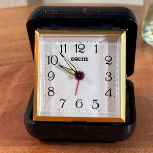 May include: A square Equity travel alarm clock with a white face, black numerals, and gold-tone trim. The clock is housed in a black hinged case that is open, revealing the clock face with black hour and minute hands and a thin red second hand.