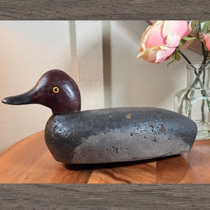 May include: A wooden duck decoy, hand-carved with a dark burgundy head and black beak. The body is painted in shades of grey and brown. The duck is on a wooden surface. A vase of pink flowers is in the background.
