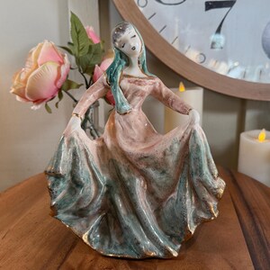 May include: A vintage porcelain figurine of a woman in a flowing dress. The dress is primarily pink and teal with gold trim. The woman has her eyes closed and is holding the dress out. The figurine is on a wooden surface.