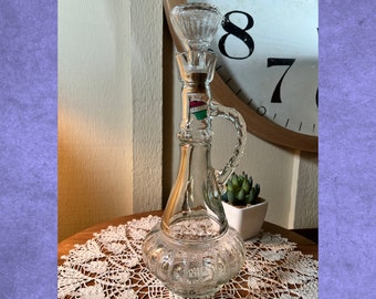 Op de afbeelding: Een karaf van helder glas met een getextureerde, bolvormige basis en een bijpassende glazen stop. De karaf heeft een gebogen handvat met een decoratief patroon en een klein rechthoekig label op de hals met onleesbare tekst. Het glas heeft een geperst, gefacetteerd ontwerp over het hele lichaam en de stop, wat zorgt voor een reflecterend oppervlak.
