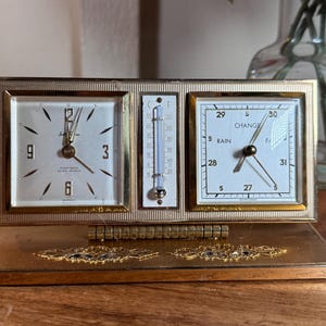 May include: A rectangular gold-tone desk clock featuring a white clock face with gold numerals, a central thermometer with Celsius and Fahrenheit scales, and a barometer dial labeled with Rain, Change, and Fair. The base has decorative gold accents.