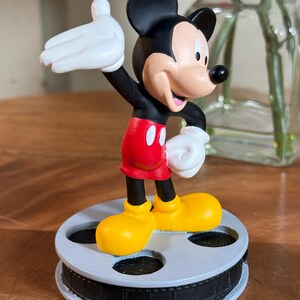 May include: A Mickey Mouse figurine in a classic pose, wearing red shorts, yellow shoes, and white gloves. The black-eared character stands on a film reel base. The figurine is a collectible item.