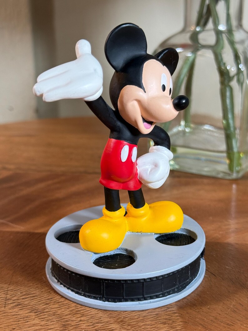 May include: A Mickey Mouse figurine in a classic pose, wearing red shorts, yellow shoes, and white gloves. The black-eared character stands on a film reel base. The figurine is a collectible item.