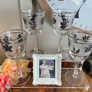 May include: Four frosted glass wine glasses with silver leaf accents in the shape of leaves. The glasses are arranged on a tiered acrylic display stand. A small white picture frame with a black and white photo of a man in a suit is in the center of the stand.