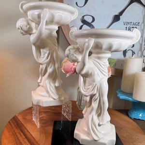 May include: Two cream-coloured ceramic sculptures of figures holding decorative bowls. The figures are in a classical style, with draped clothing. The bowls have ornate handles. The sculptures are on a wooden surface.