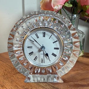 Puede incluir: Un reloj de cuarzo de cristal transparente con esfera redonda y números romanos. El reloj tiene un borde decorativo festoneado y se apoya sobre una superficie de madera. La esfera es blanca con números y agujas negras.