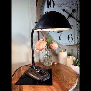 May include: A black desk lamp with a flexible neck and a dome-shaped shade. The lamp sits on a black square base. The base is on a wooden table. A clock and flowers are in the background.