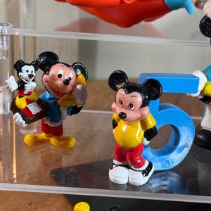 May include: A collection of Mickey Mouse figurines in various poses and outfits. The figurines are brightly colored, featuring black, red, yellow, and blue. One figurine is standing next to a blue number 5. An orange and blue toy airplane is visible in the background.