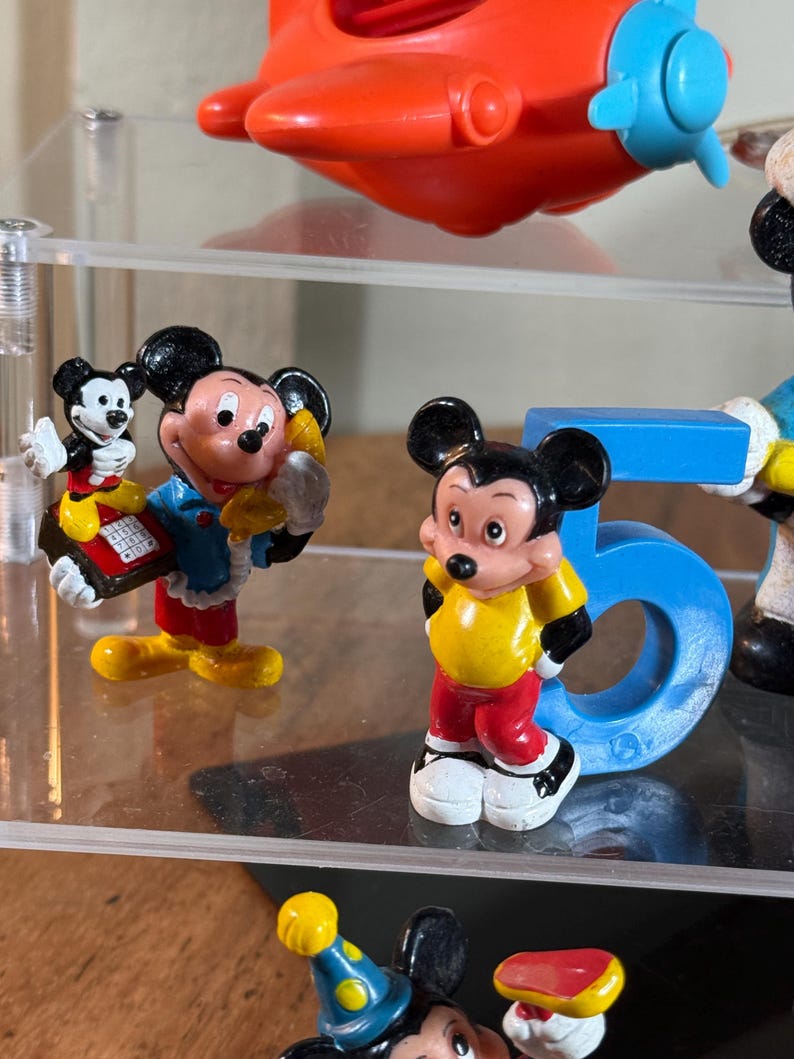May include: A collection of Mickey Mouse figurines in various poses and outfits. The figurines are brightly colored, featuring black, red, yellow, and blue. One figurine is standing next to a blue number 5. An orange and blue toy airplane is visible in the background.