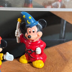 May include: A Mickey Mouse figurine dressed as a sorcerer, wearing a blue hat with yellow stars and a crescent moon. He is in a red robe and yellow shoes, holding a white wand with a yellow star. The figurine is a collectible item.