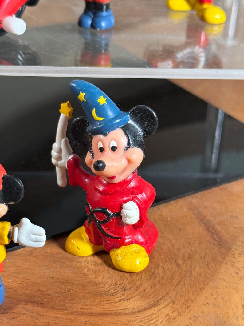 May include: A Mickey Mouse figurine dressed as a sorcerer, wearing a blue hat with yellow stars and a crescent moon. He is in a red robe and yellow shoes, holding a white wand with a yellow star. The figurine is a collectible item.