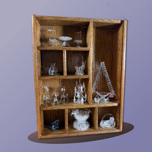 May include: A wooden display shelf filled with miniature glass figurines. The shelf has multiple compartments, each holding a different glass object, including animals, furniture, and decorative pieces. The shelf is made of wood with a natural finish.