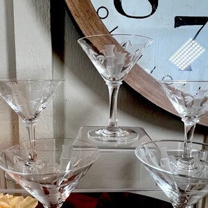 Op de afbeelding: Vier heldere glazen martini glazen met gegraveerde ontwerpen op een transparant acryl oppervlak. De glazen zijn in een rij gerangschikt, met de twee aan de uiteinden lichtjes gekanteld naar het midden.