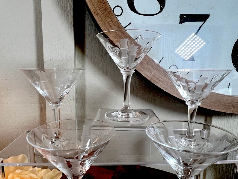 Op de afbeelding: Vier heldere glazen martini glazen met gegraveerde ontwerpen op een transparant acryl oppervlak. De glazen zijn in een rij gerangschikt, met de twee aan de uiteinden lichtjes gekanteld naar het midden.