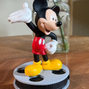 May include: A Mickey Mouse figurine in a classic pose, wearing red shorts, yellow shoes, and white gloves. The black and white figure stands on a film reel base. The background includes a glass vase with flowers.