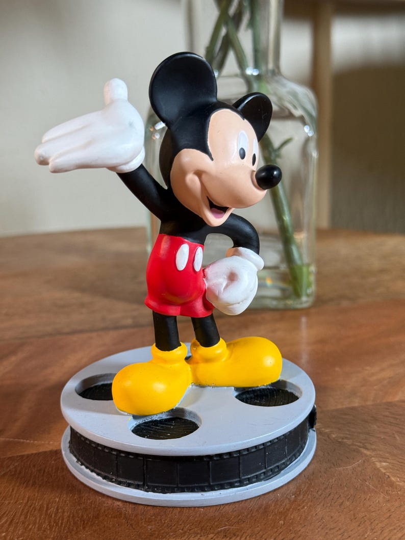 May include: A Mickey Mouse figurine in a classic pose, wearing red shorts, yellow shoes, and white gloves. The black and white figure stands on a film reel base. The background includes a glass vase with flowers.