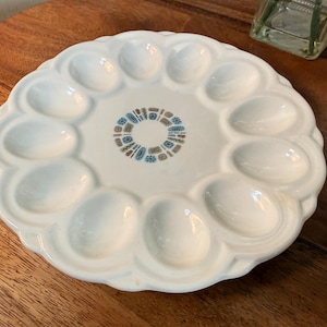 May include: White ceramic egg serving platter with 12 egg-shaped indentations. The center features a circular design with blue, brown, and tan geometric shapes. The platter has a scalloped edge and is on a wooden surface.