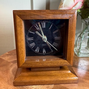 May include: A square wooden clock with a dark face and Roman numerals. The clock has a rectangular base and a wooden frame. The clock hands are silver. The clock is sitting on a wooden surface.