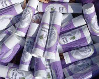 May include: A close-up of a pile of purple and white lip balm tubes. The tubes are labeled "Very Vanilla Lip Balm" and "Wanna Do Herbs!" with a drawing of a woman and lavender flowers.