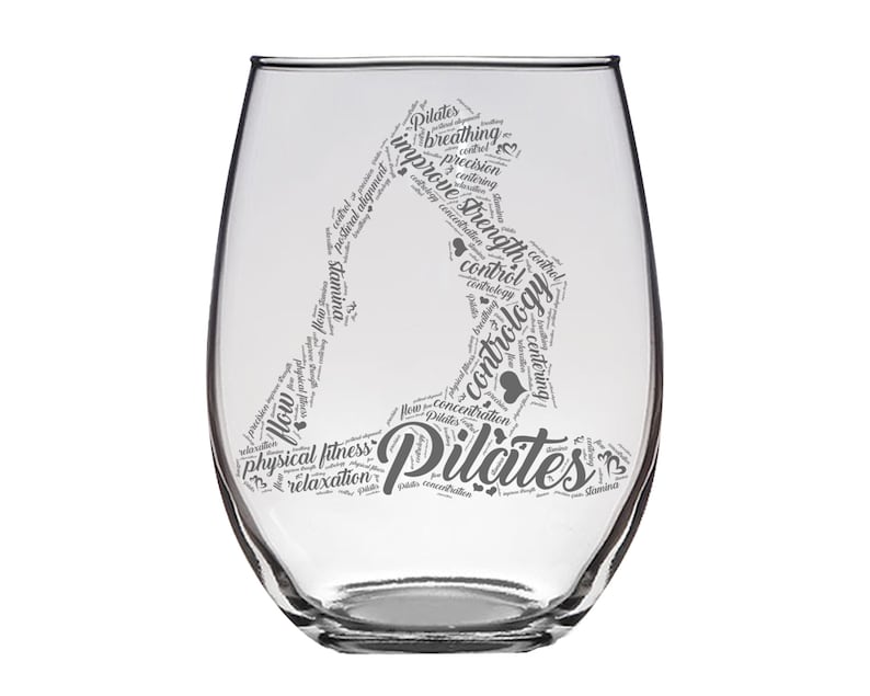 May include: A clear glass wine glass with a design of a person doing a Pilates pose. The design is made up of words related to Pilates, such as "breathing", "control", "strength", and "concentration".