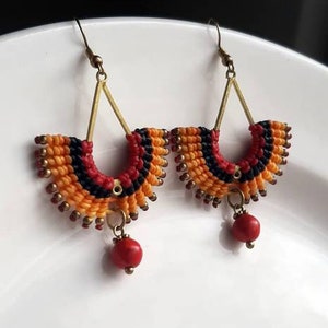 May include: A pair of handmade earrings with a fan-shaped design. The earrings are made with orange, red, and black thread and have a small red bead dangling from the bottom.